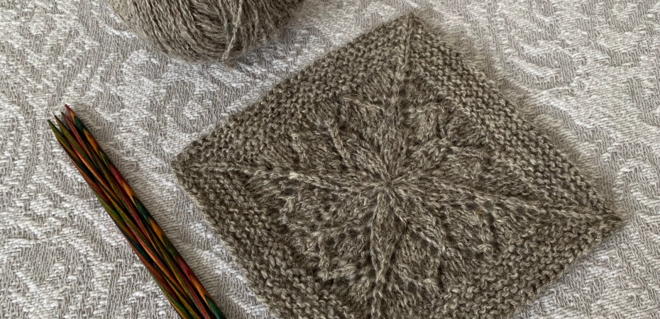 A grey wool blanket square with lace motif in the centre sits next to a small ball of grey wool and a set of double pointed needles.