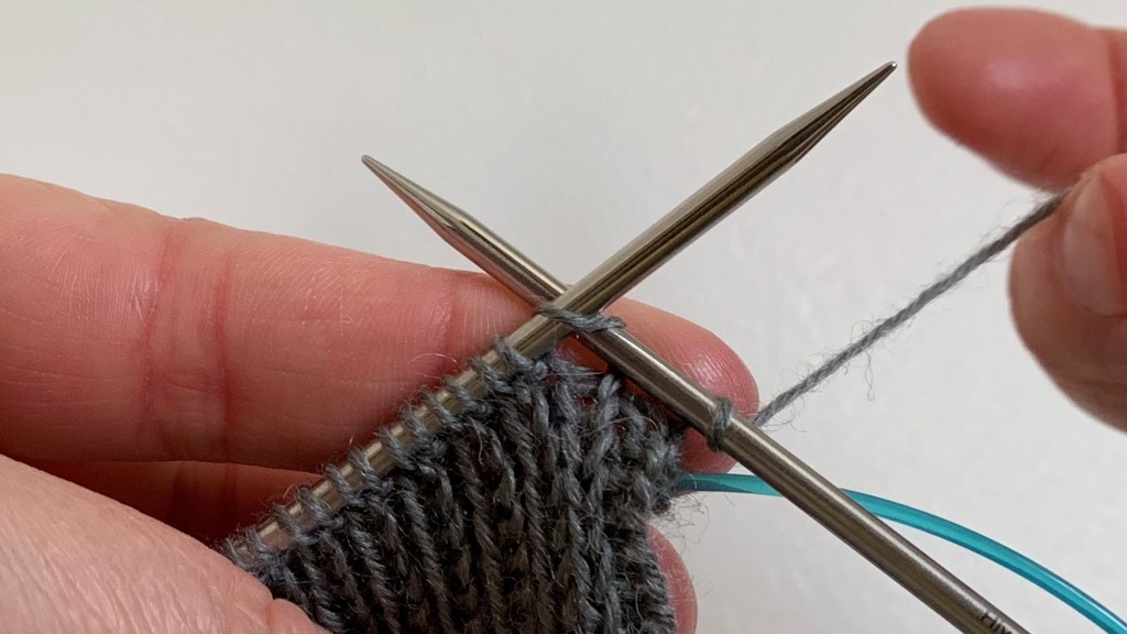 Stretchy bind off. Setting up for the first stitch. Working yarn is carried over and under the right needle which is inserted in to the first stitch on the left needle ready to knit.