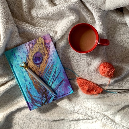 Image shows a pen sat on a peacock feather journal, to the right is a mug of tea and the beginnings of a toe-up sock